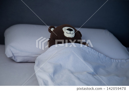 Top view of a bed with gray linens in which lies a brown teddy bear, covered with a blanket, his head resting on a soft pillow. The concept of a cozy sleep, a children's bedroom. High quality photo Top view of a bed with gray linens in which lies a brown teddy bear, covered with a blanket, his head resting on a soft pillow. The concept of a cozy sleep, a children's bedroom. High quality photo 142059478