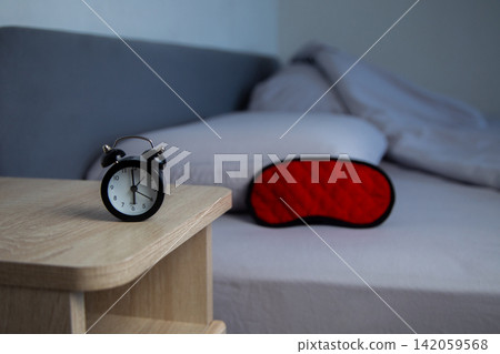 In the foreground is a wooden bedside table with a black alarm clock, in the background is a blurred gray bed with a bright red sleep mask. The concept of early awakening, healthy sleep, sleep In the foreground is a wooden bedside table with a black alarm clock, in the background is a blurred gray bed with a bright red sleep mask. The concept of early awakening, healthy sleep, sleep 142059568