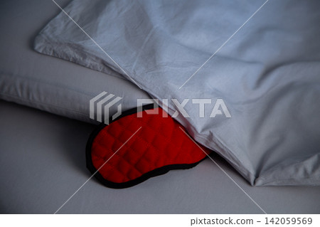 A close-up of a red sleep mask covered with a blanket, leaning against a pillow and inviting to sleep against a background of gray pastel linen. The concept of healthy sleep, sleep patterns, and A close-up of a red sleep mask covered with a blanket, leaning against a pillow and inviting to sleep against a background of gray pastel linen. The concept of healthy sleep, sleep patterns, and 142059569