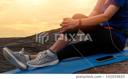 Asian woman resting after a beach workout at sunrise while wearing a smart fitness tracker and earphones. Recovery, mindfulness, healthy lifestyle, wearable technology and tropical coastal wellness. Asian woman resting after a beach workout at sunrise while wearing a smart fitness tracker and earphones. Recovery, mindfulness, healthy lifestyle, wearable technology and tropical coastal wellness. 142059685