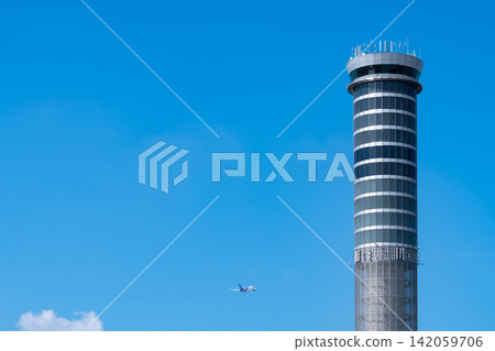Airport air traffic control tower with commercial airplane flying under clear blue sky. Aviation technology, flight management, civil aviation infrastructure and global transportation network. Airport air traffic control tower with commercial airplane flying under clear blue sky. Aviation technology, flight management, civil aviation infrastructure and global transportation network. 142059706