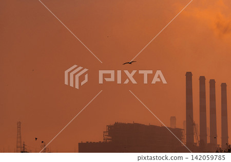 Industrial power plant with smokestacks silhouetted against sunrise sky. Electricity generation, energy infrastructure, atmospheric haze, environmental impact and global industrial development. Industrial power plant with smokestacks silhouetted against sunrise sky. Electricity generation, energy infrastructure, atmospheric haze, environmental impact and global industrial development. 142059785