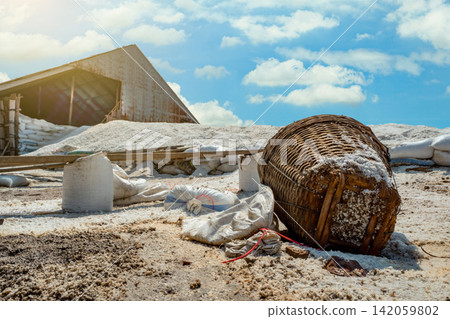 Traditional sea salt production with bamboo basket, raw organic salt piles and storage barn under blue summer sky, natural mineral harvesting process for food industry and manufacturing. Traditional sea salt production with bamboo basket, raw organic salt piles and storage barn under blue summer sky, natural mineral harvesting process for food industry and manufacturing. 142059802