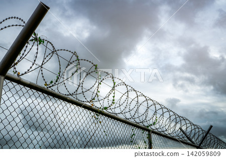 Razor wire security fence beneath dramatic storm clouds, concept of border security, military defense, prison protection, restricted access, surveillance systems and critical infrastructure safety. Razor wire security fence beneath dramatic storm clouds, concept of border security, military defense, prison protection, restricted access, surveillance systems and critical infrastructure safety. 142059809