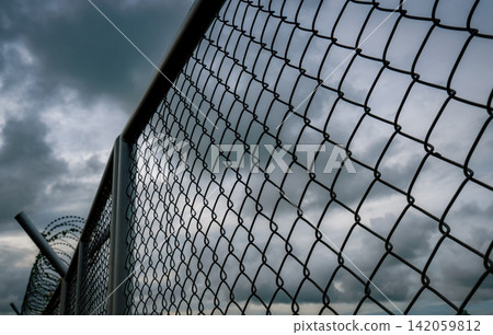 Chain link security fence with razor wire beneath dramatic storm clouds. Military protection, border security, restricted access, prison perimeter, surveillance and critical infrastructure defense. 142059812