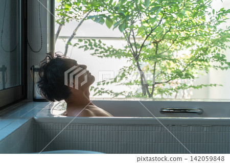 A middle-aged Asian man soaking in a bathtub in his home bathroom (bath time, bathing, bath). A middle-aged Asian man soaking in a bathtub in his home bathroom (bath time, bathing, bath). 142059848