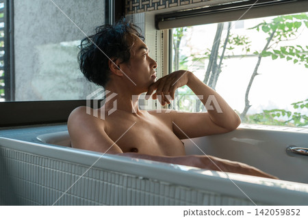 A middle-aged Asian man soaking in a bathtub in his home bathroom (bath time, bathing, bath). A middle-aged Asian man soaking in a bathtub in his home bathroom (bath time, bathing, bath). 142059852