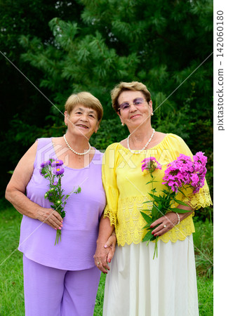 Two happy Russian senior sisters hold flowers and stand together during outdoor activities. Two happy Russian senior sisters hold flowers and stand together during outdoor activities. 142060180
