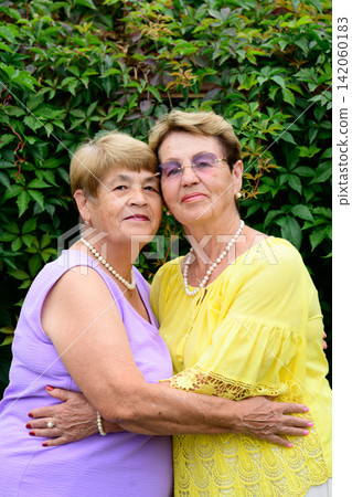 Two happy Russian senior sisters embracing and showing love near green ivy wall at rural garden. Two happy Russian senior sisters embracing and showing love near green ivy wall at rural garden. 142060183