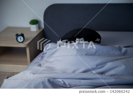 Top view on a gray bed under a blanket lies a brown teddy bear with a black sleep mask, next to a bedside table there is a mini alarm clock and a white pot with a plant. The concept of a sleeping Top view on a gray bed under a blanket lies a brown teddy bear with a black sleep mask, next to a bedside table there is a mini alarm clock and a white pot with a plant. The concept of a sleeping 142060242