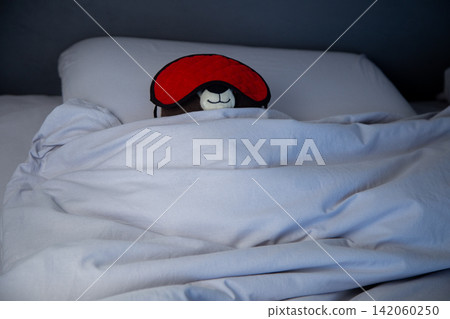 Close-up of a brown teddy bear lying on a pillow, covered with a blanket, with a red sleep mask on a gray pastel background. The concept of early awakening, healthy sleep, sleep patterns, and the Close-up of a brown teddy bear lying on a pillow, covered with a blanket, with a red sleep mask on a gray pastel background. The concept of early awakening, healthy sleep, sleep patterns, and the 142060250