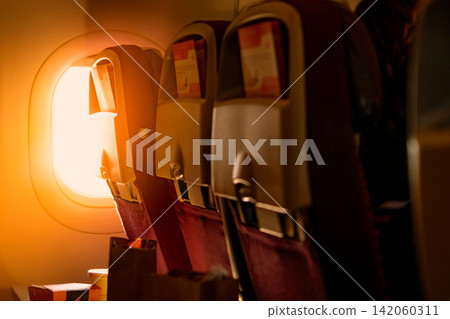 Warm golden sunlight streaming through airplane window illuminating passenger seats inside commercial aircraft cabin. Comfortable air travel, airline hospitality, tourism, vacation and modern aviation Warm golden sunlight streaming through airplane window illuminating passenger seats inside commercial aircraft cabin. Comfortable air travel, airline hospitality, tourism, vacation and modern aviation 142060311