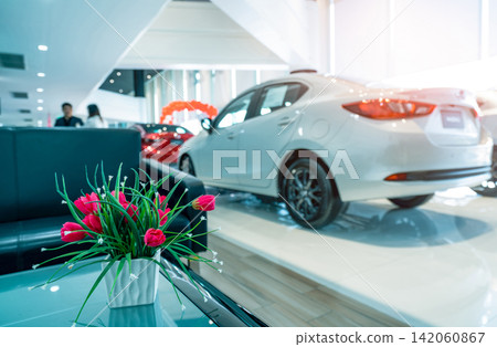 Fake red flowers in ceramic white vase on blurred background of sale person deals with customer in modern showroom. Car dealership. Blurred luxury car parked in showroom. Automotive business industry. Fake red flowers in ceramic white vase on blurred background of sale person deals with customer in modern showroom. Car dealership. Blurred luxury car parked in showroom. Automotive business industry. 142060867