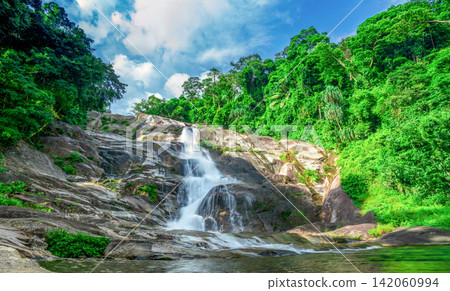 Beautiful tropical waterfall cascading over granite mountain rocks beneath blue sky. Lush rainforest in Thailand. Wilderness landscape for travel and nature theme. Ecotourism and conservation. Beautiful tropical waterfall cascading over granite mountain rocks beneath blue sky. Lush rainforest in Thailand. Wilderness landscape for travel and nature theme. Ecotourism and conservation. 142060994