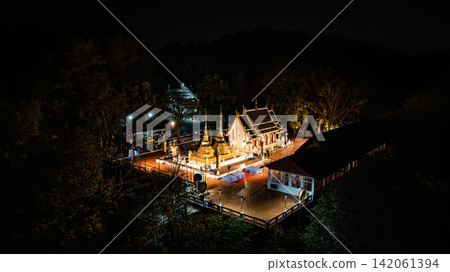 Phra That Doi Tung rises above the dark highland landscape, its radiant golden architecture contrasting with the forest and distant lights in a serene aerial scene from 2023. 142061394