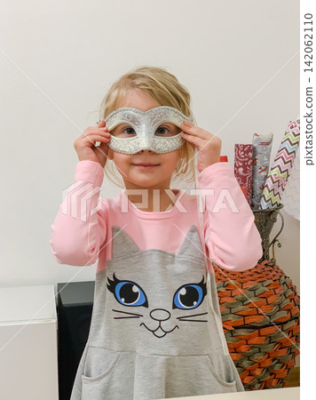 A little girl wearing a white carnival cat mask standing indoors in a cozy room A little girl wearing a white carnival cat mask standing indoors in a cozy room 142062110
