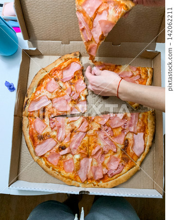 Top view of a person reaching for a slice of ham pizza from an open cardboard takeaway box on a table at home while enjoying a meal Top view of a person reaching for a slice of ham pizza from an open cardboard takeaway box on a table at home while enjoying a meal 142062211
