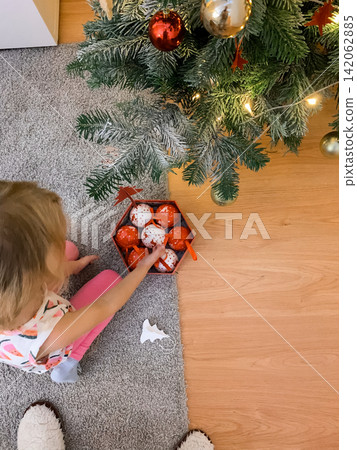 Young girl sitting on the carpet and taking red Christmas baubles from a box to decorate the tree Young girl sitting on the carpet and taking red Christmas baubles from a box to decorate the tree 142062885