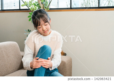 A girl in elementary and middle school sitting on a sofa at home holding her knees and shins due to knee pain A girl in elementary and middle school sitting on a sofa at home holding her knees and shins due to knee pain 142063421
