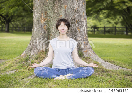 一位年輕女子盤腿坐在大樹前;這是戶外瑜珈的畫面。 一位年輕女子盤腿坐在大樹前;這是戶外瑜珈的畫面。 142063581