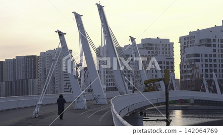 Modern Bridge Over Water At Golden Hour. Pedestrian Pathway Beside Skyscrapers Under Pastel Sky. Creative Modern Bridge Over Water At Golden Hour. Pedestrian Pathway Beside Skyscrapers Under Pastel Sky. Creative 142064769