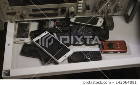 Old mobile phones stacked in white drawer beneath vintage electronic equipment, showing worn plastic casings, buttons, screens, cables, and industrial control details in cluttered workspace 142064881