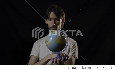 Adult man holding spinning tabletop globe in dark studio, gesturing toward viewer with focused expression, blue meridian stand, glossy surface, centered front view, educational concept 142064944