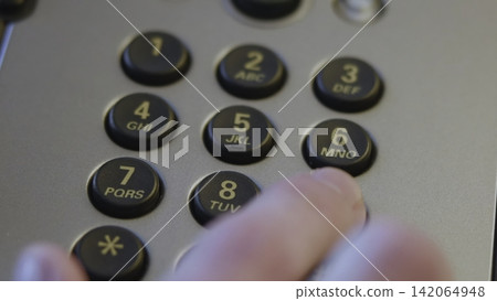 Close-up fingertip pressing numeric keypad on office desk telephone under warm diffuse light, showing tactile contact, recessed round keys, matte silver housing, shallow focus 142064948