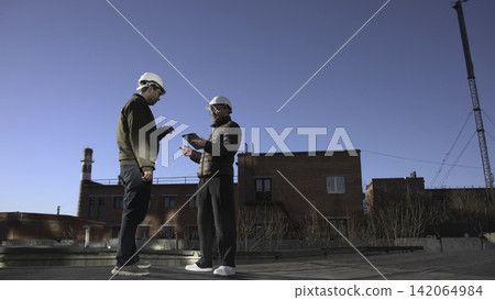 Group Of Construction Laborers Carefully Analyzing Blueprints On Rooftop Amidst Clear Sky Conditions. Media Group Of Construction Laborers Carefully Analyzing Blueprints On Rooftop Amidst Clear Sky Conditions. Media 142064984