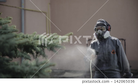 Person in protective hooded coverall spraying fine mist toward evergreen tree in urban courtyard beside pastel wall with safety gear, soft daylight, and visible moisture Person in protective hooded coverall spraying fine mist toward evergreen tree in urban courtyard beside pastel wall with safety gear, soft daylight, and visible moisture 142065036