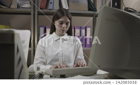 Young woman typing at office desk in storage style back office with binders, beige keyboard, desktop computer, and CRT monitor visible in tidy clerical workspace Young woman typing at office desk in storage style back office with binders, beige keyboard, desktop computer, and CRT monitor visible in tidy clerical workspace 142065038