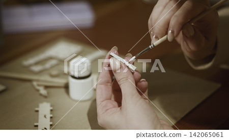 Hands applying white paint onto thin wooden model strip with fine brush during indoor craft session, precise edge work and manicure detail on steady tabletop, with hand, painting, wooden, brush detail Hands applying white paint onto thin wooden model strip with fine brush during indoor craft session, precise edge work and manicure detail on steady tabletop, with hand, painting, wooden, brush detail 142065061