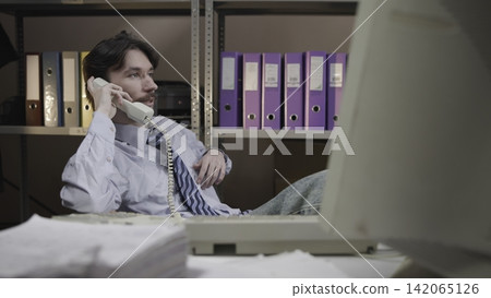 Young office worker seated at desk listening on corded telephone with colorful binders on metal shelving behind him Young office worker seated at desk listening on corded telephone with colorful binders on metal shelving behind him 142065126