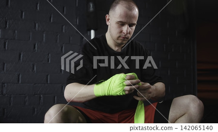 Male athlete wrapping neon green hand wraps before boxing workout inside dim gym with dark brick wall, focused posture, strong contrast, and gritty preparation mood Male athlete wrapping neon green hand wraps before boxing workout inside dim gym with dark brick wall, focused posture, strong contrast, and gritty preparation mood 142065140