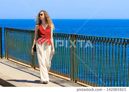 A young woman strolls along a scenic coastal path, enjoying the ocean view and summer vacation atmosphere A young woman strolls along a scenic coastal path, enjoying the ocean view and summer vacation atmosphere 142065923