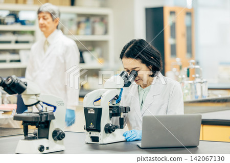 一位五十多歲的女研究員,身穿白色實驗服,正在透過顯微鏡觀察。 一位五十多歲的女研究員,身穿白色實驗服,正在透過顯微鏡觀察。 142067130