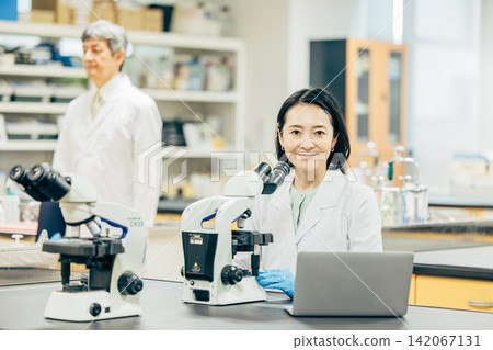 一位五十多歲的女研究員,身穿白色實驗服,正在透過顯微鏡觀察。 一位五十多歲的女研究員,身穿白色實驗服,正在透過顯微鏡觀察。 142067131