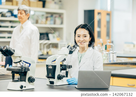 一位五十多歲的女研究員,身穿白色實驗服,正在透過顯微鏡觀察。 一位五十多歲的女研究員,身穿白色實驗服,正在透過顯微鏡觀察。 142067132