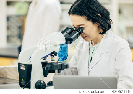 A female researcher in her 50s, wearing a white lab coat, peers through a microscope. A female researcher in her 50s, wearing a white lab coat, peers through a microscope. 142067136