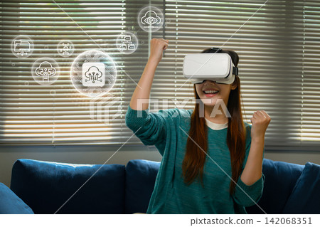 Excited Asian Woman Wearing VR Headset with Floating Cloud Technology Icons in Living Room Excited Asian Woman Wearing VR Headset with Floating Cloud Technology Icons in Living Room 142068351