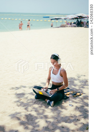 Side view of fit woman using laptop while sitting on the sand at a tropical beach Side view of fit woman using laptop while sitting on the sand at a tropical beach 142068380