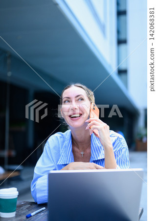 Smiling woman in blue striped shirt using mobile phone and laptop at a resort 142068381