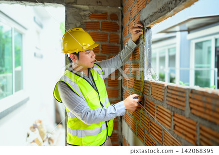 Asian Engineer in Yellow Hard Hat Measuring Brick Wall at Construction Site 142068395