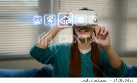 Young Asian Woman Wearing VR Headset Touching Glowing Cloud Computing Icons in Virtual Reality Interface Young Asian Woman Wearing VR Headset Touching Glowing Cloud Computing Icons in Virtual Reality Interface 142068452