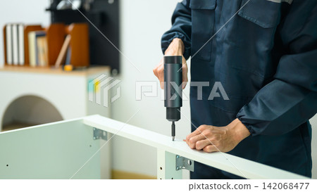 Technician in Blue Uniform Using Electric Screwdriver to Assemble White Furniture Cabinet Indoors 142068477