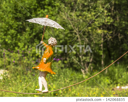 Whimsical Garden Statue Balancing on Tightrope Holding Lace Umbrella in Sunny Green Park Whimsical Garden Statue Balancing on Tightrope Holding Lace Umbrella in Sunny Green Park 142068835