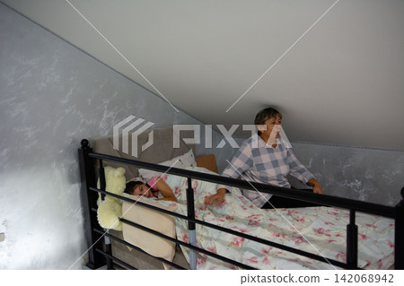 Grandmother watching over granddaughter sleeping in attic bedroom with sloped ceiling and floral bedding in subdued colors. Grandmother watching over granddaughter sleeping in attic bedroom with sloped ceiling and floral bedding in subdued colors. 142068942
