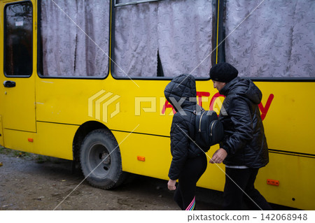 People boarding yellow bus. Quilted coats and a backpack. Curtained windows. Suggests travel or evacuation in a somber setting. People boarding yellow bus. Quilted coats and a backpack. Curtained windows. Suggests travel or evacuation in a somber setting. 142068948