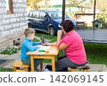 A young boy and his grandmother are drawing outdoors together at a small table, enjoying a sunny day and quality time together. 142069145