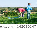 A woman lays out a blanket while a young boy, holding a tablet, watches in a grassy field near some colorful flowers on a sunny day. 142069147